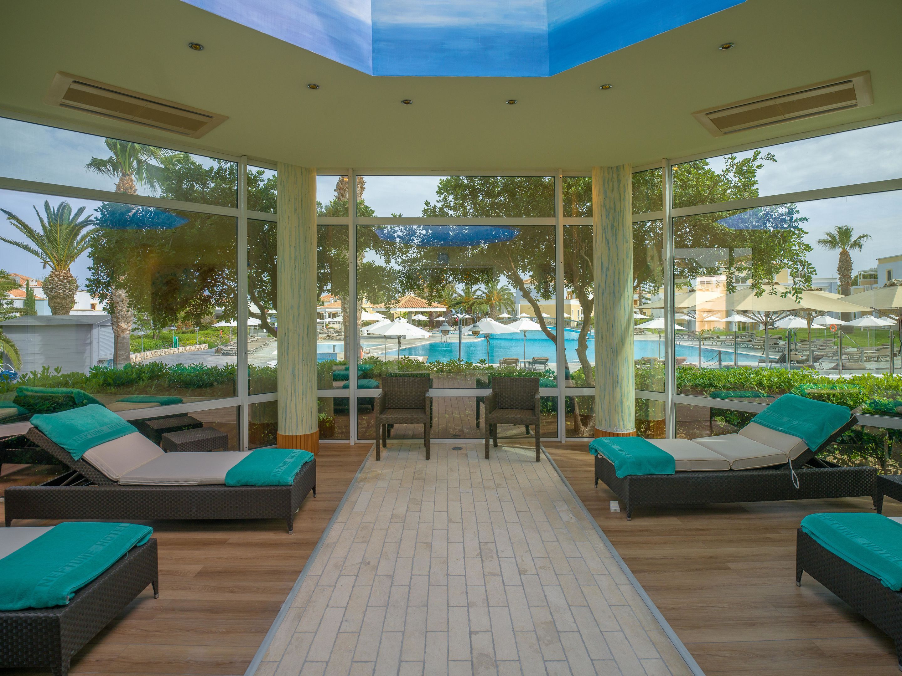 A sunlit indoor lounge area with a pool view, furnished with lounge chairs and a table.