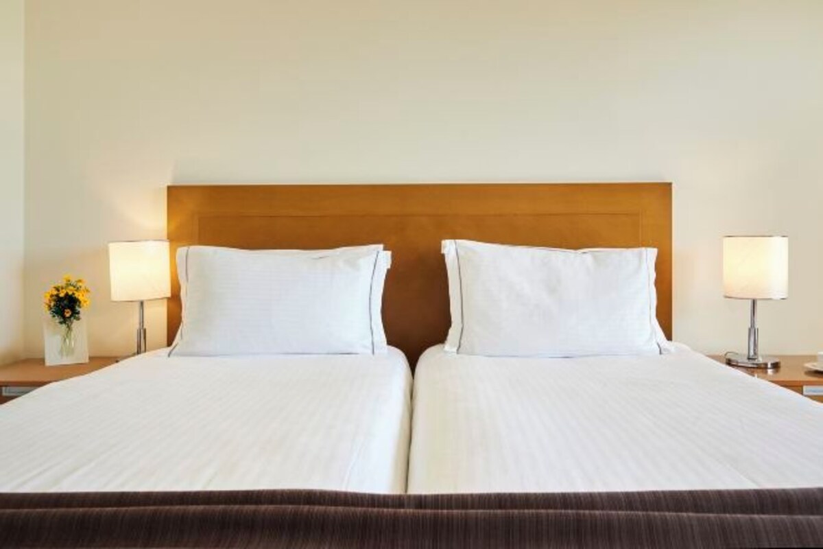 Una cama doble con cabecera de madera, dos lámparas de noche y un florero con flores en la mesita.