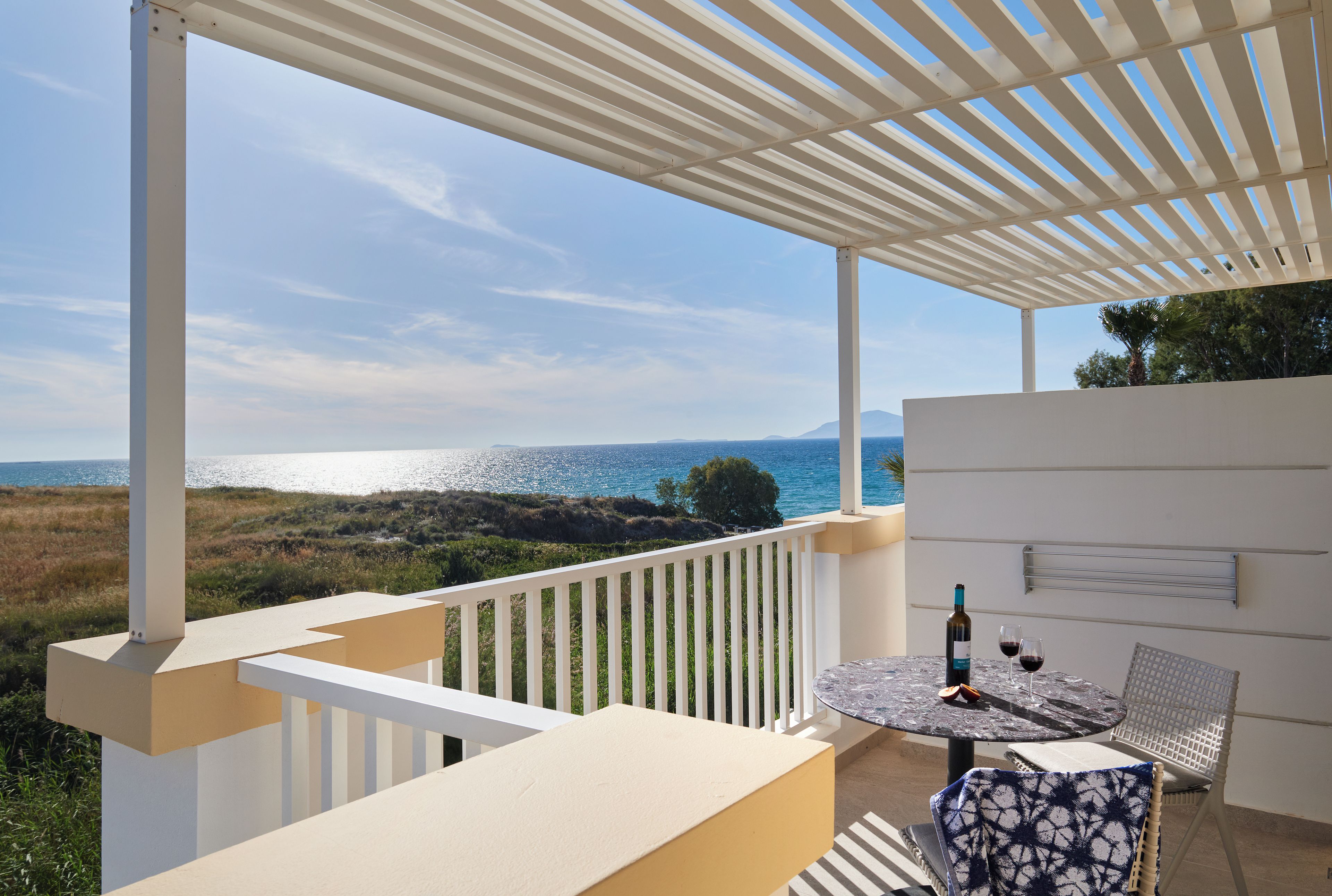 A balcony with a view of the sea, a table with a bottle and glasses, and a chair.
