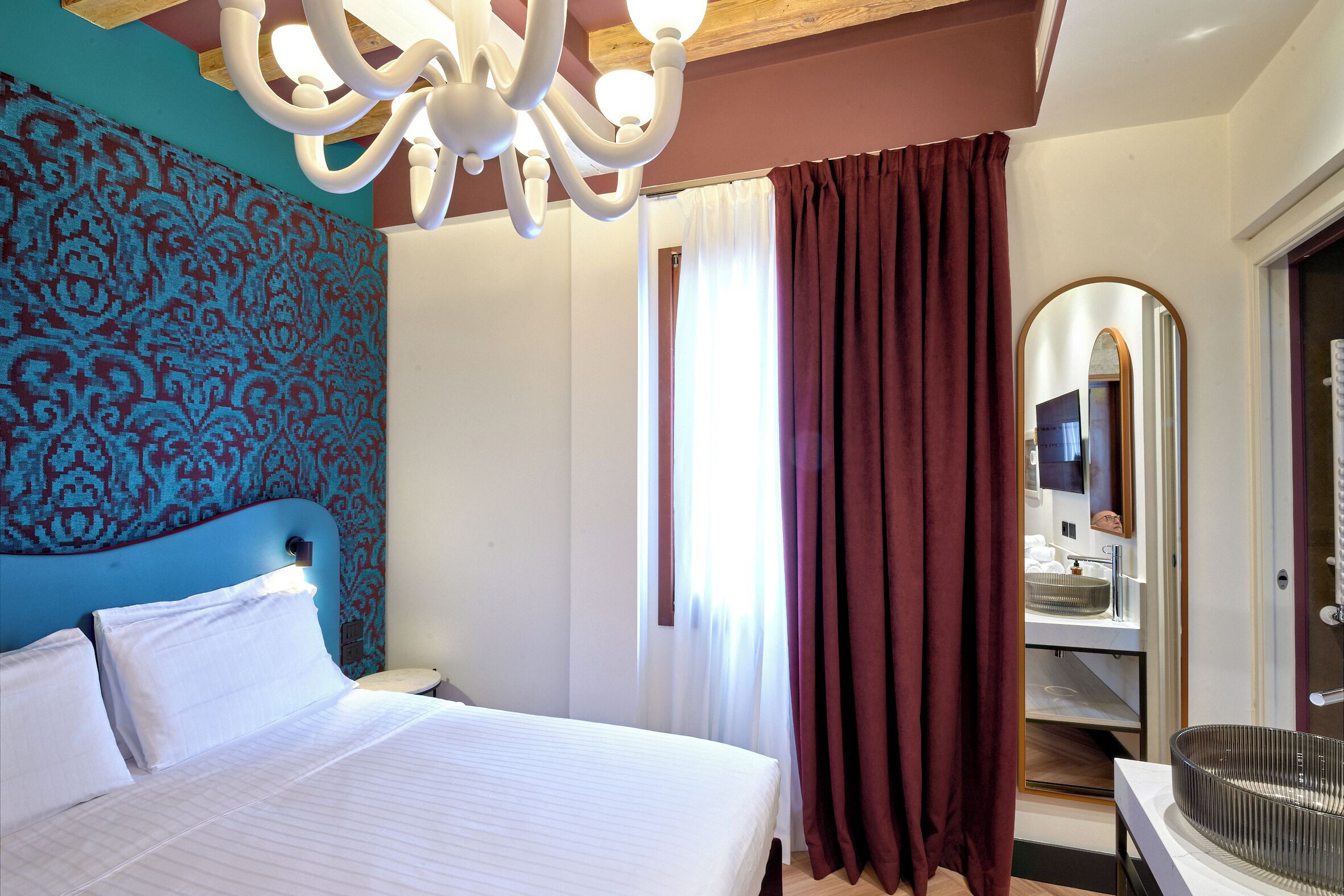 Una habitación de hotel con una cama, una araña, un espejo y un ventanal con cortinas.