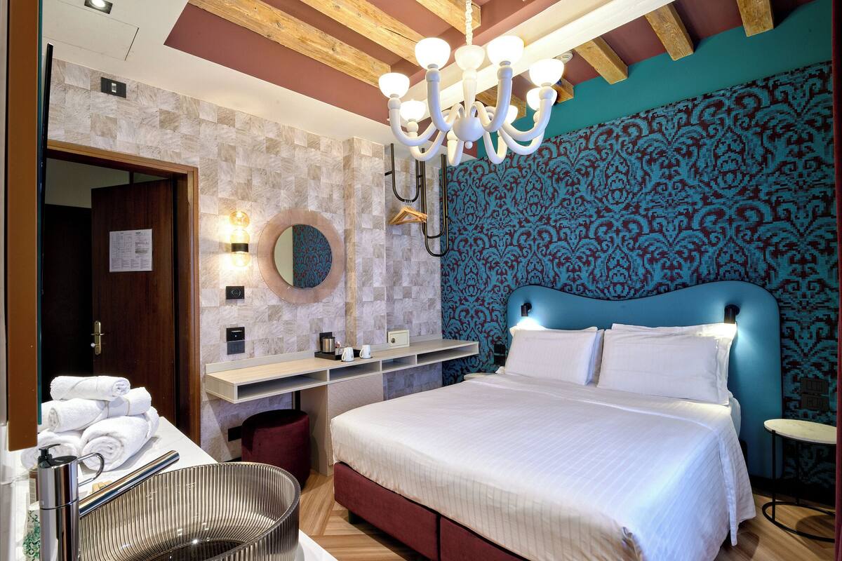 A hotel room with a bed, a vanity with a mirror, a towel rack, and a chandelier.