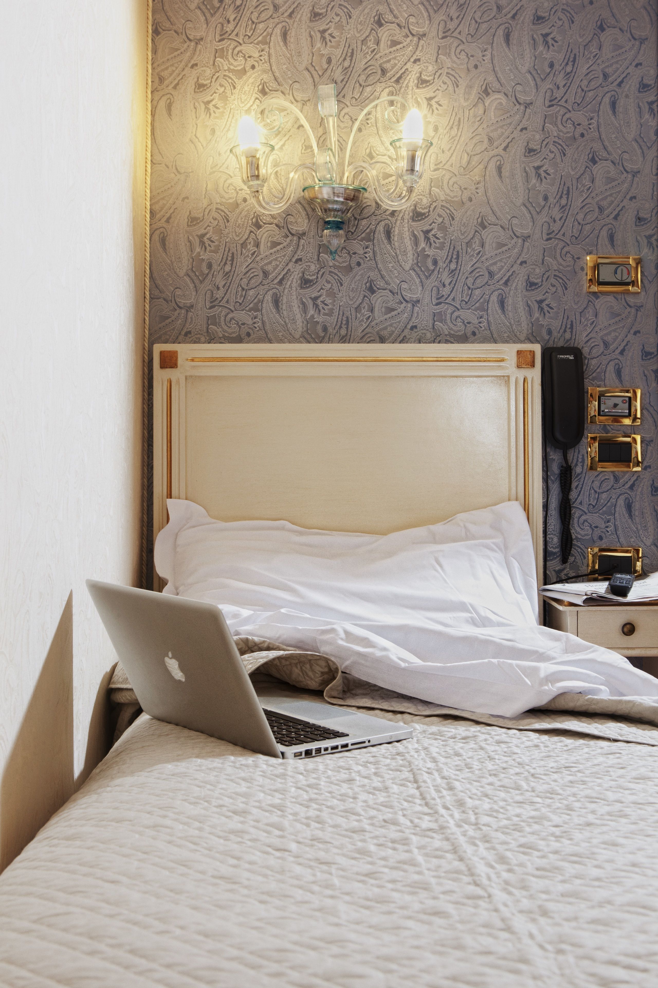 A neatly made bed with a laptop on it, a bedside table with a phone and a remote, and a wall with a patterned wallpaper and a wall-mounted light fixture.