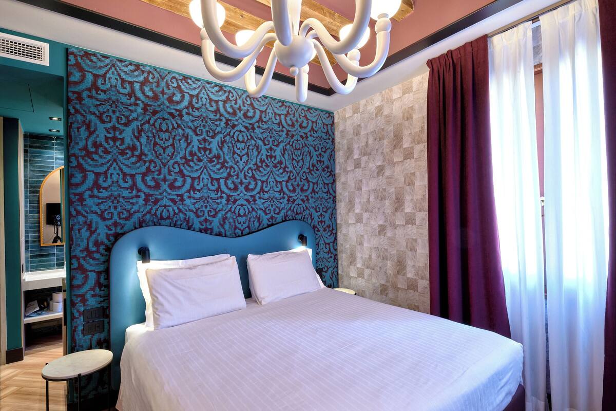 A bedroom with a large bed, a chandelier, patterned wallpaper, and curtains.