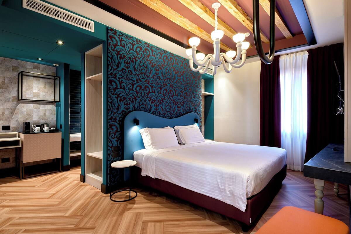 A modern hotel room with a large bed, a small round table, a built-in wardrobe, and a kitchenette.