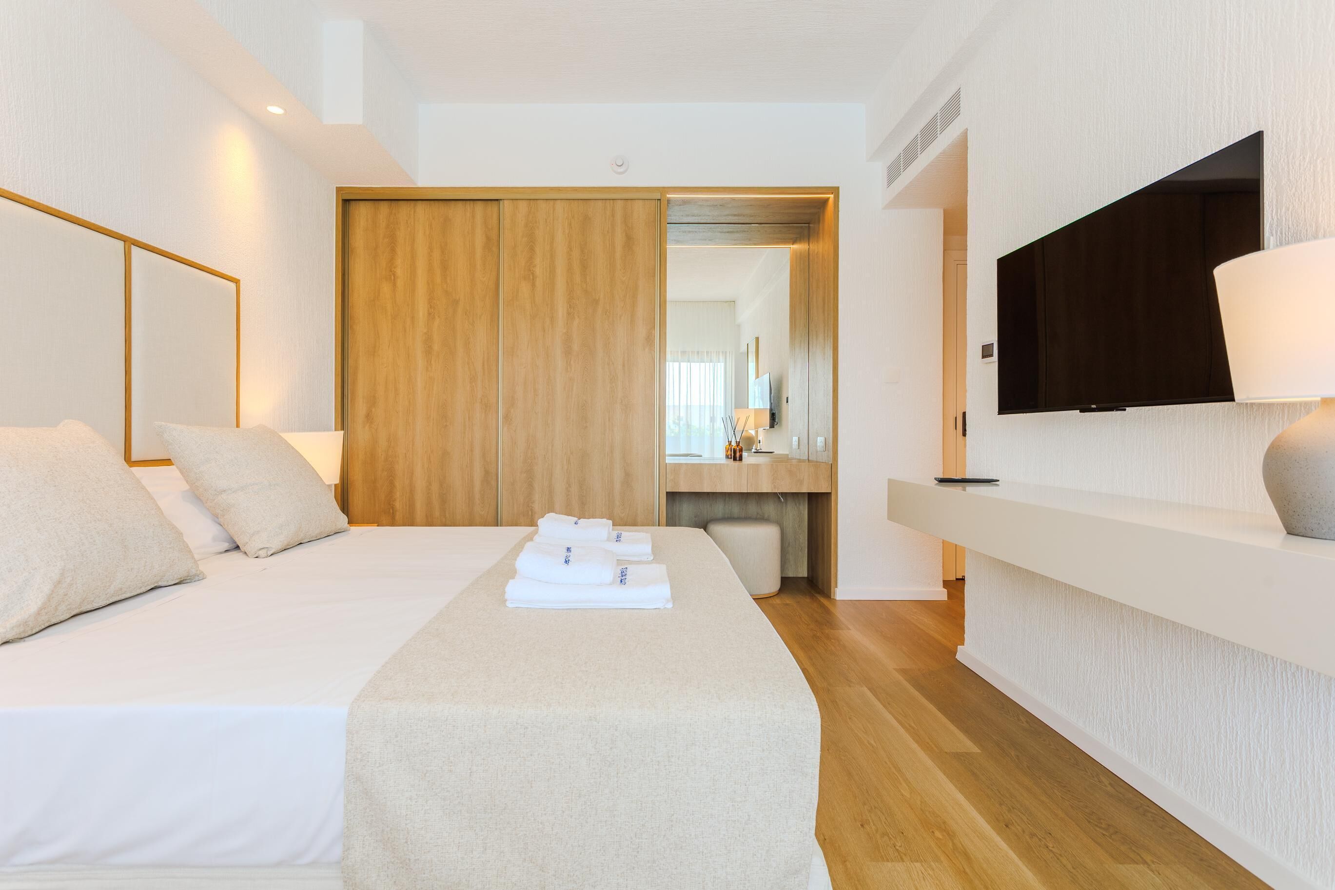 A modern hotel room with a large bed, a flat-screen TV mounted on the wall, and a wooden wardrobe.