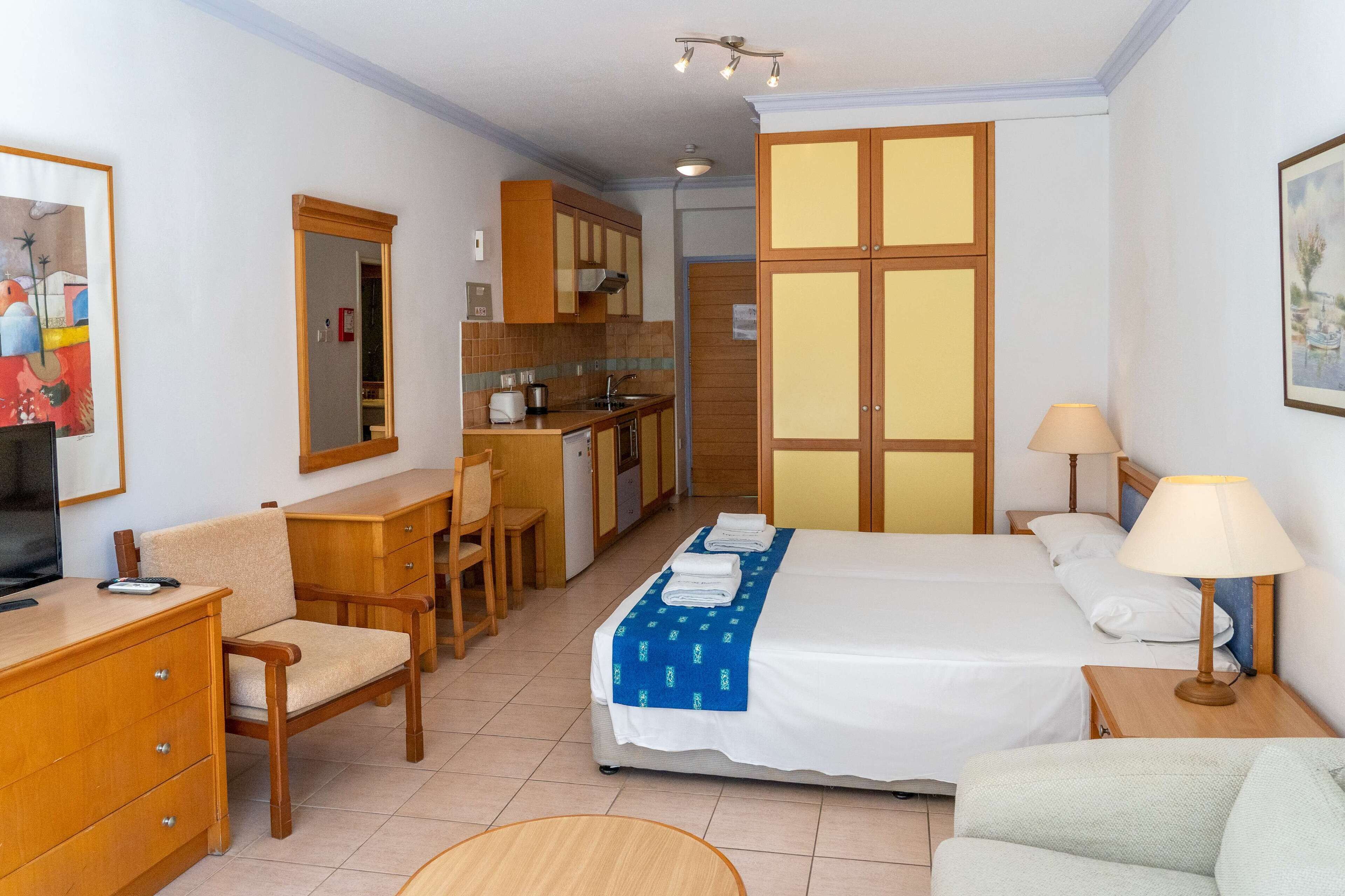 Habitación de hotel con cama, escritorio, silla, televisor, lámpara y zona de cocina.