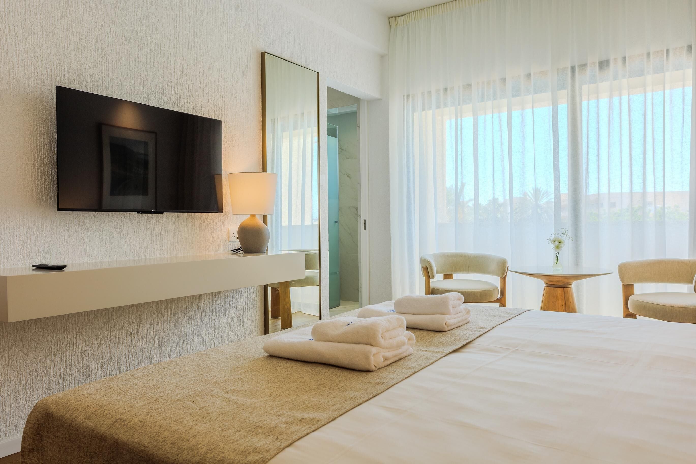 A hotel room with a bed, a TV mounted on the wall, a bedside table with a lamp, a chair, and a small table with a vase.