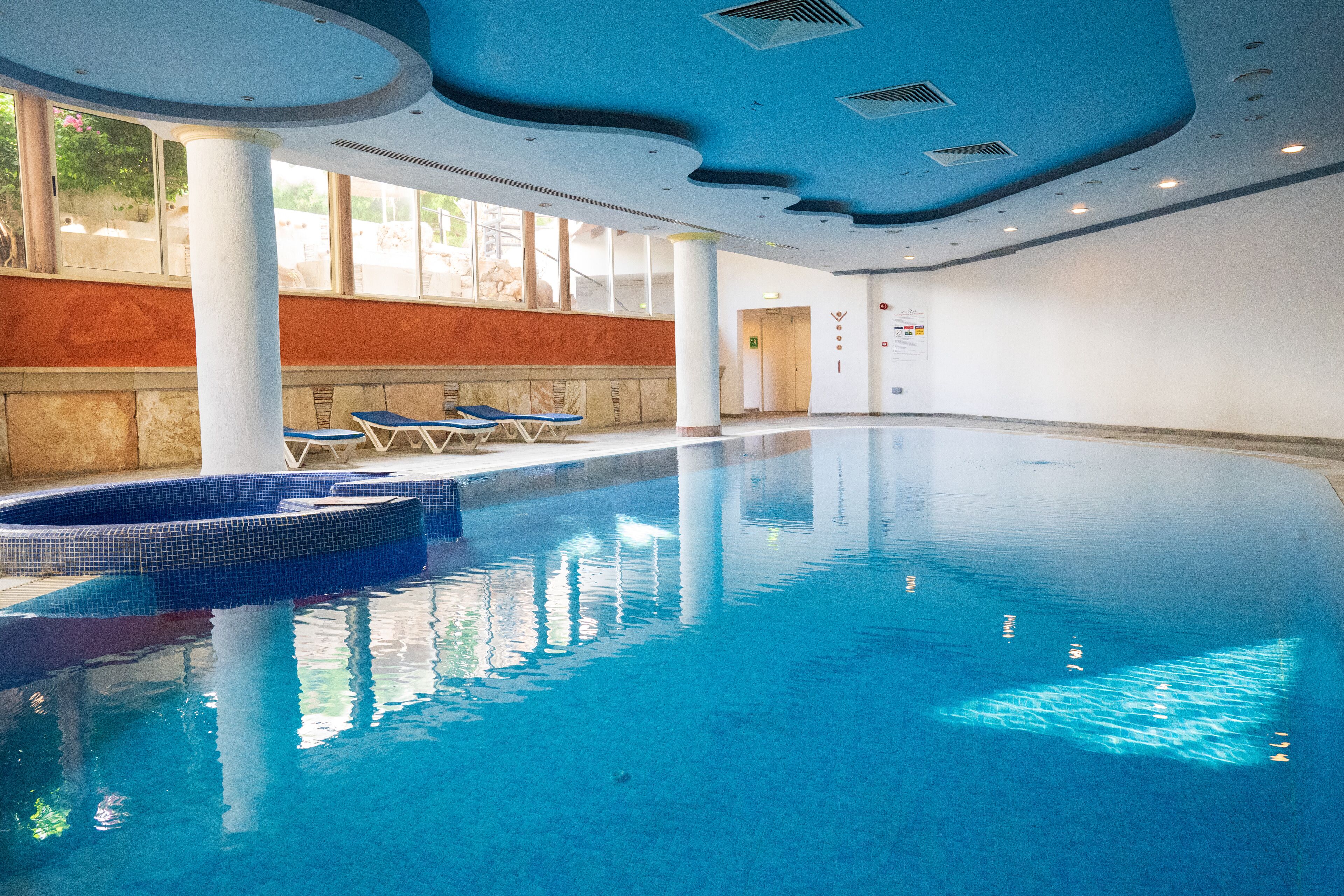 Une grande piscine intérieure à l’eau d’un bleu cristallin, entourée de murs blancs et agrémentée de quelques chaises longues.