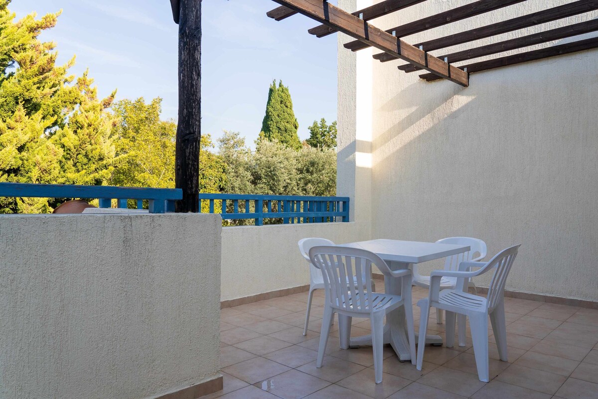 An outdoor patio with a white table and chairs, a blue railing, and a view of trees and a tall building.