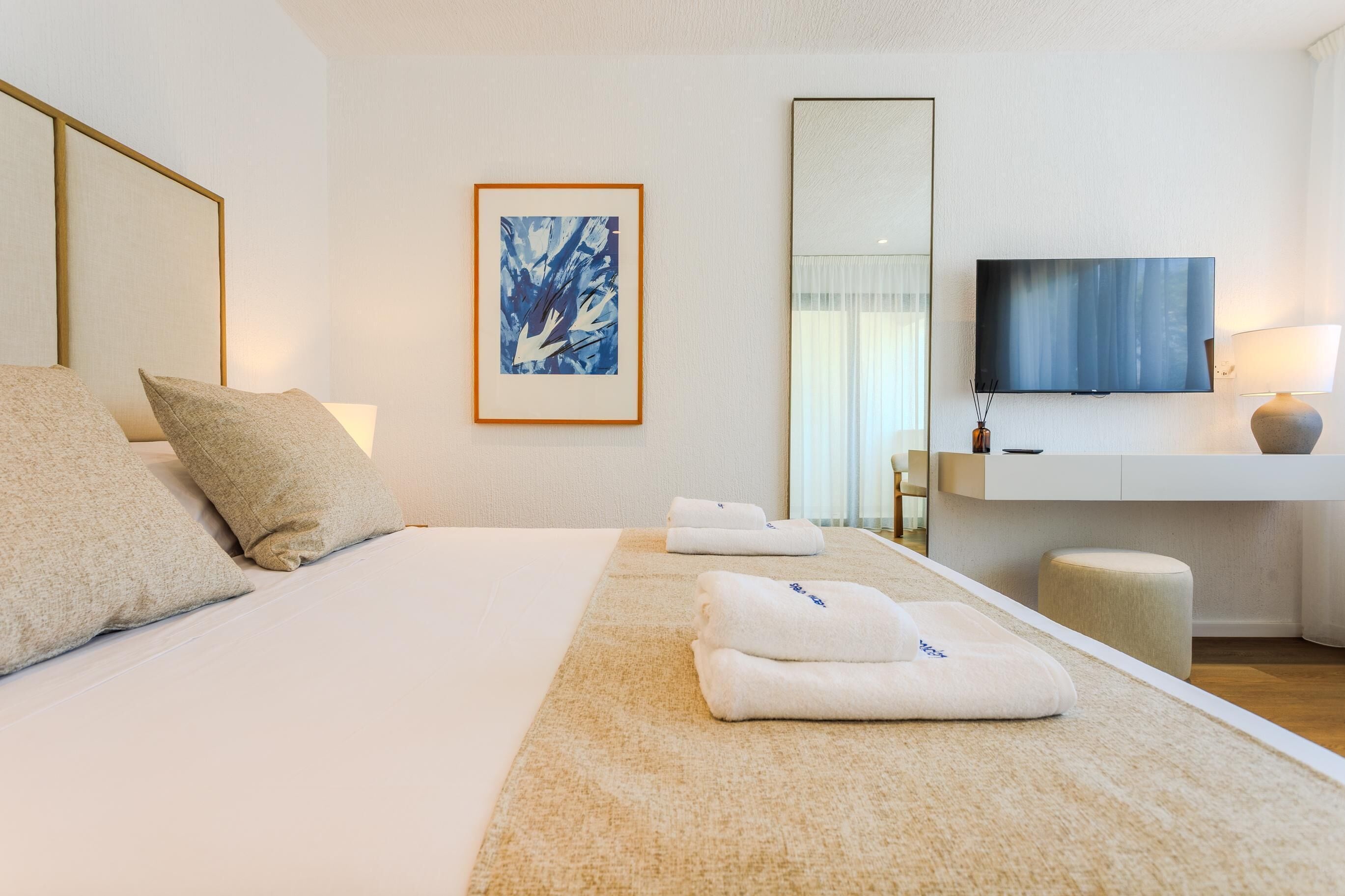 A hotel room with a bed, pillows, a framed artwork, a television, and a bedside table with a lamp.