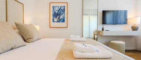 Quarto de hotel com cama, almofadas, uma obra de arte emoldurada, televisão e mesa de cabeceira com abajur.