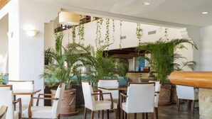 Un restaurante con sillas y mesas blancas, plantas en macetas y un área de cocina al fondo.