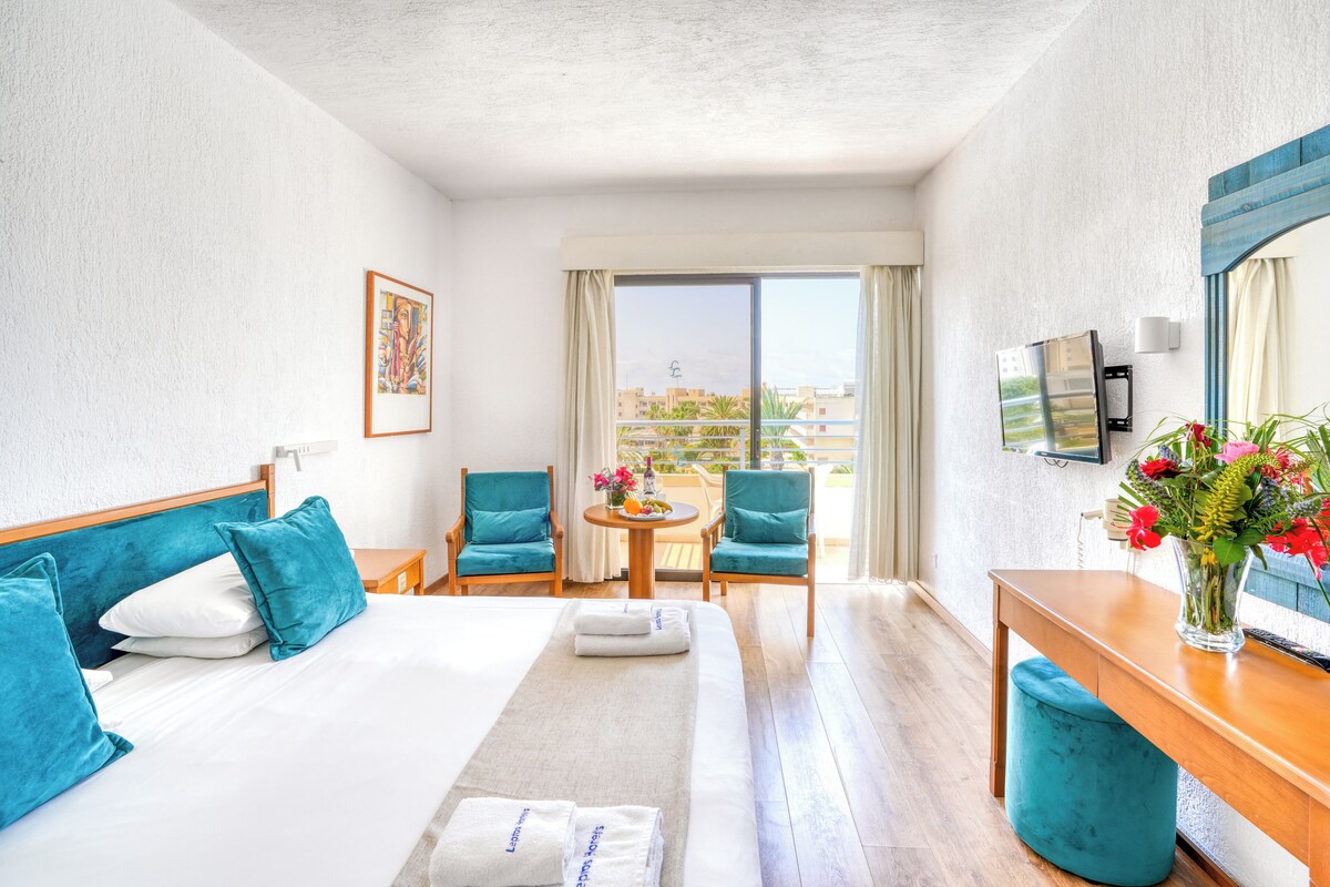 A hotel room with a bed, two chairs, a small table with flowers, a TV, and a balcony with a view.