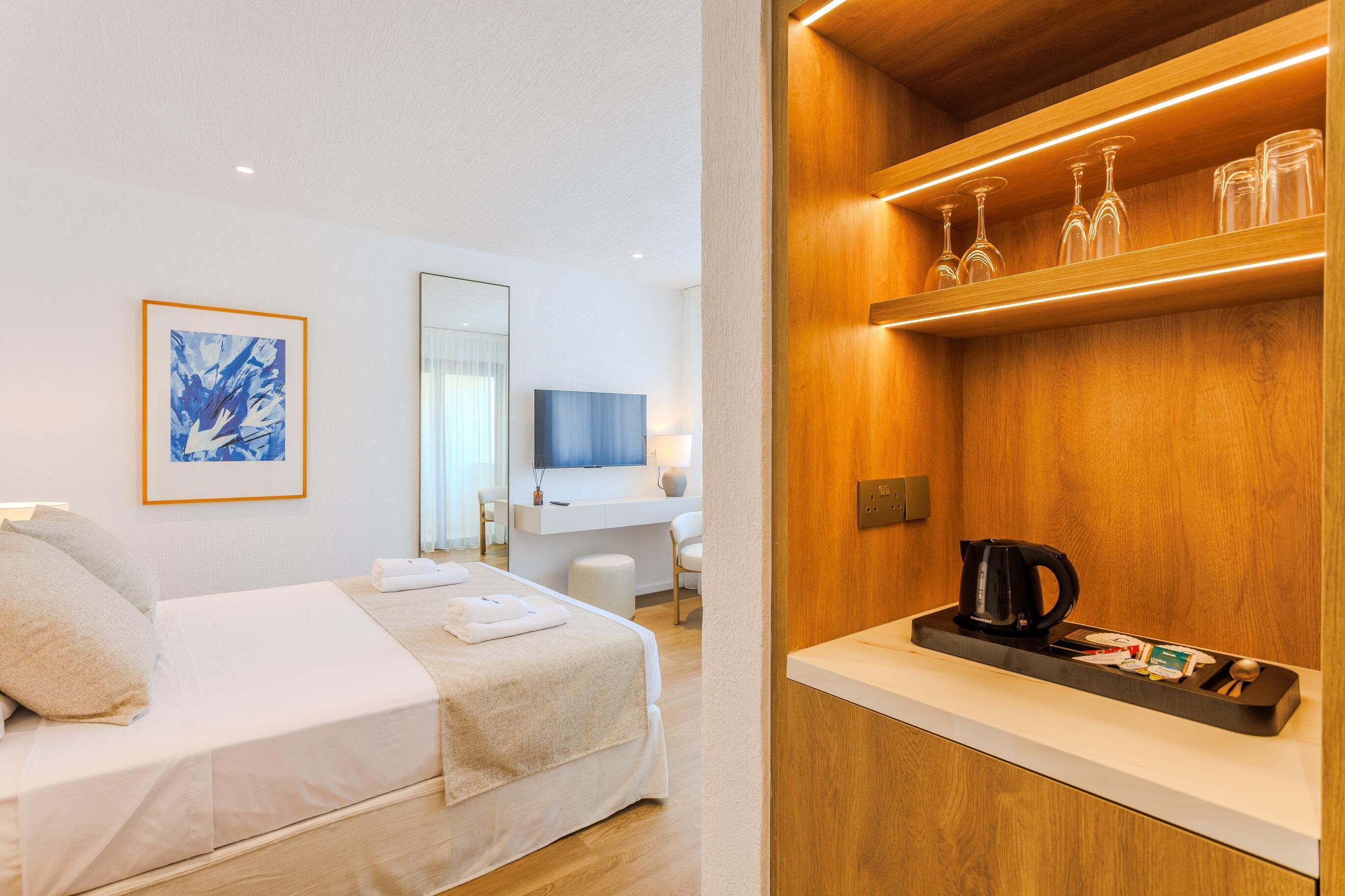 A hotel room with a bed, a bedside table with a kettle, a shelf with glasses, a TV, and a painting on the wall.