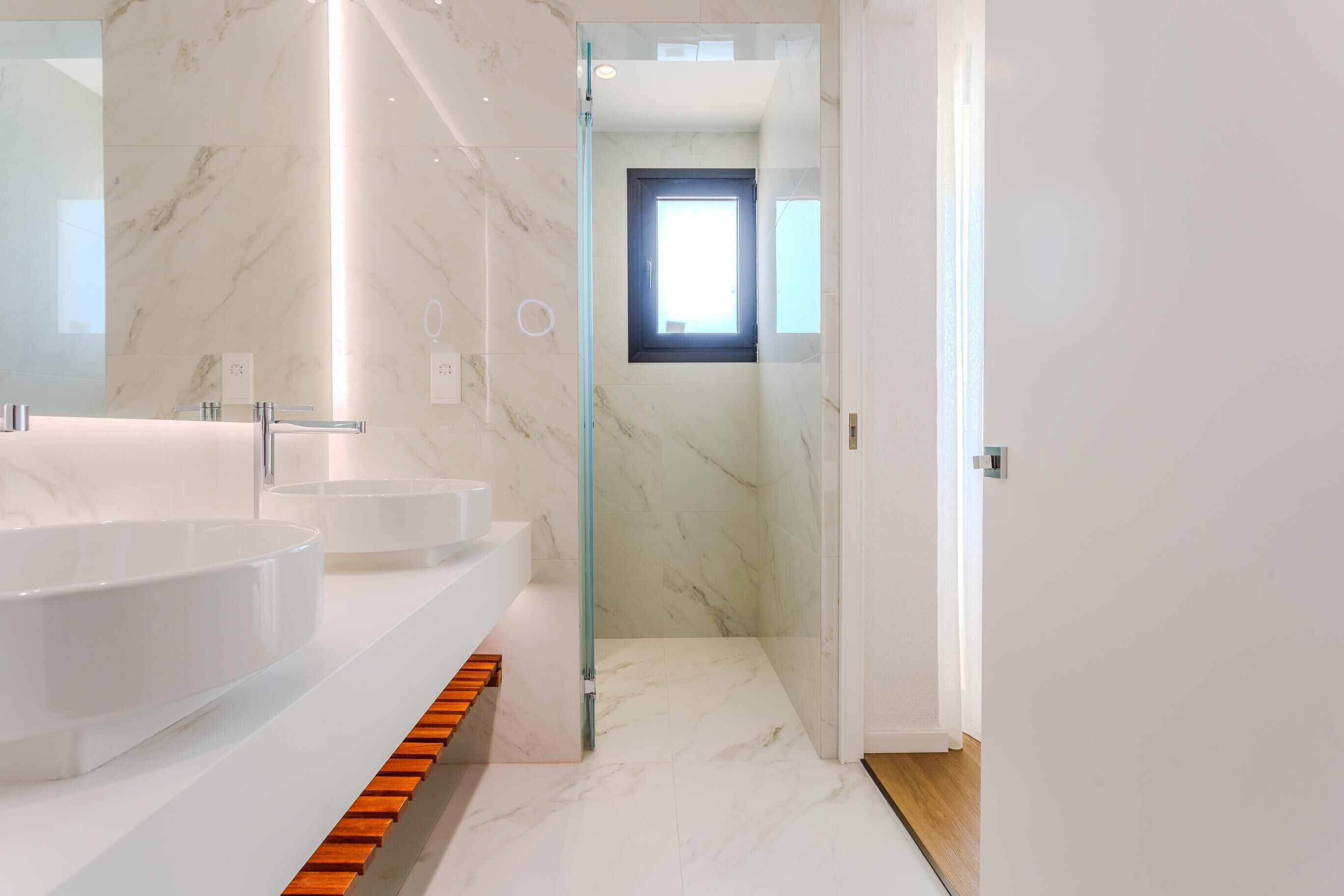 A modern bathroom with two sinks, a glass shower enclosure, and a wooden step leading to a door.