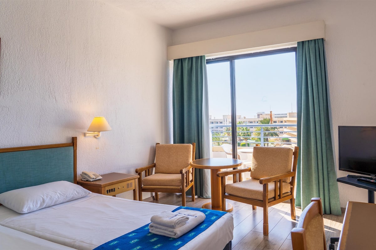A hotel room with a bed, two chairs, a small table, a TV, and a balcony with curtains.