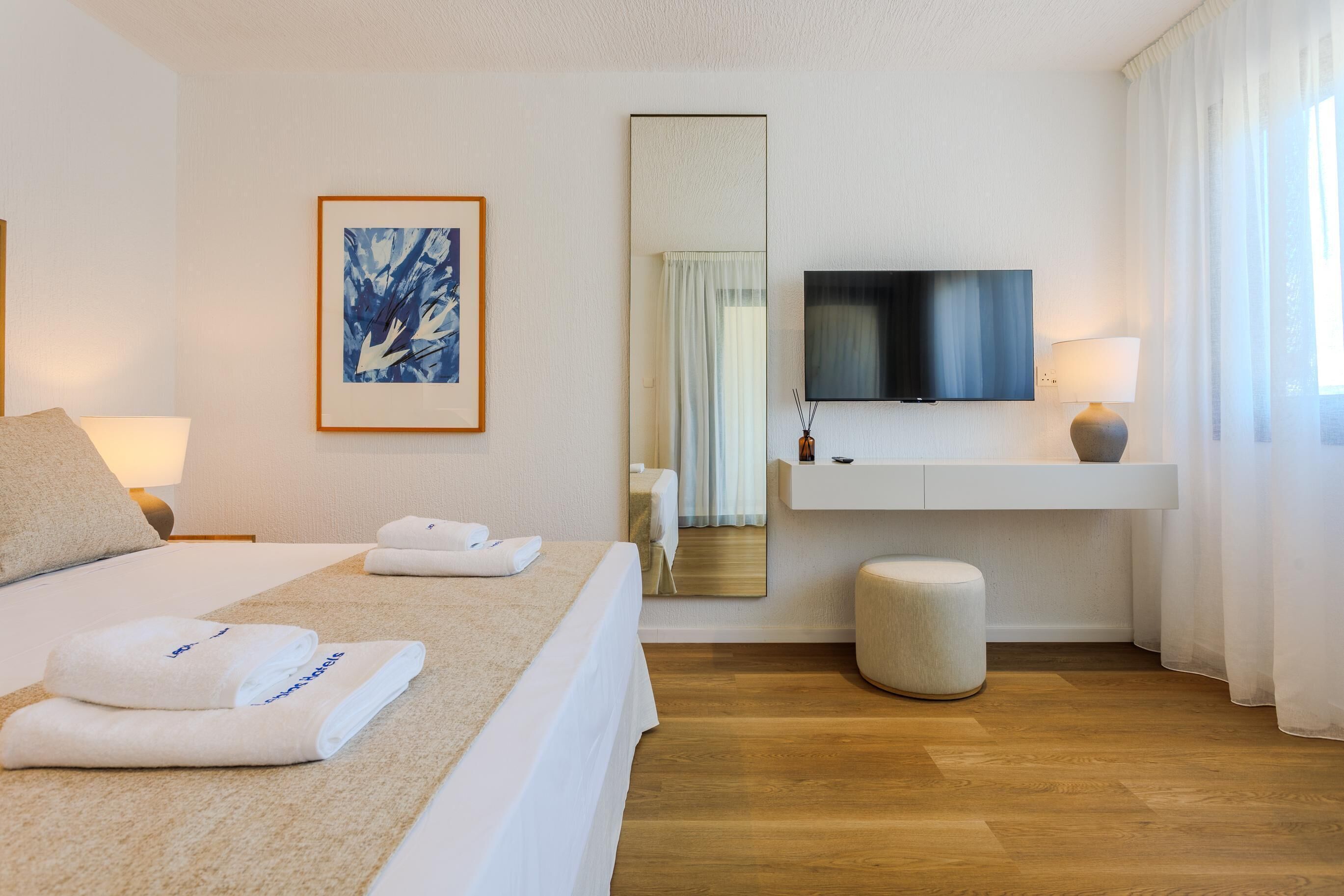 A hotel room with a bed, a flat-screen TV, a painting, a mirror, and a bedside table with a lamp.