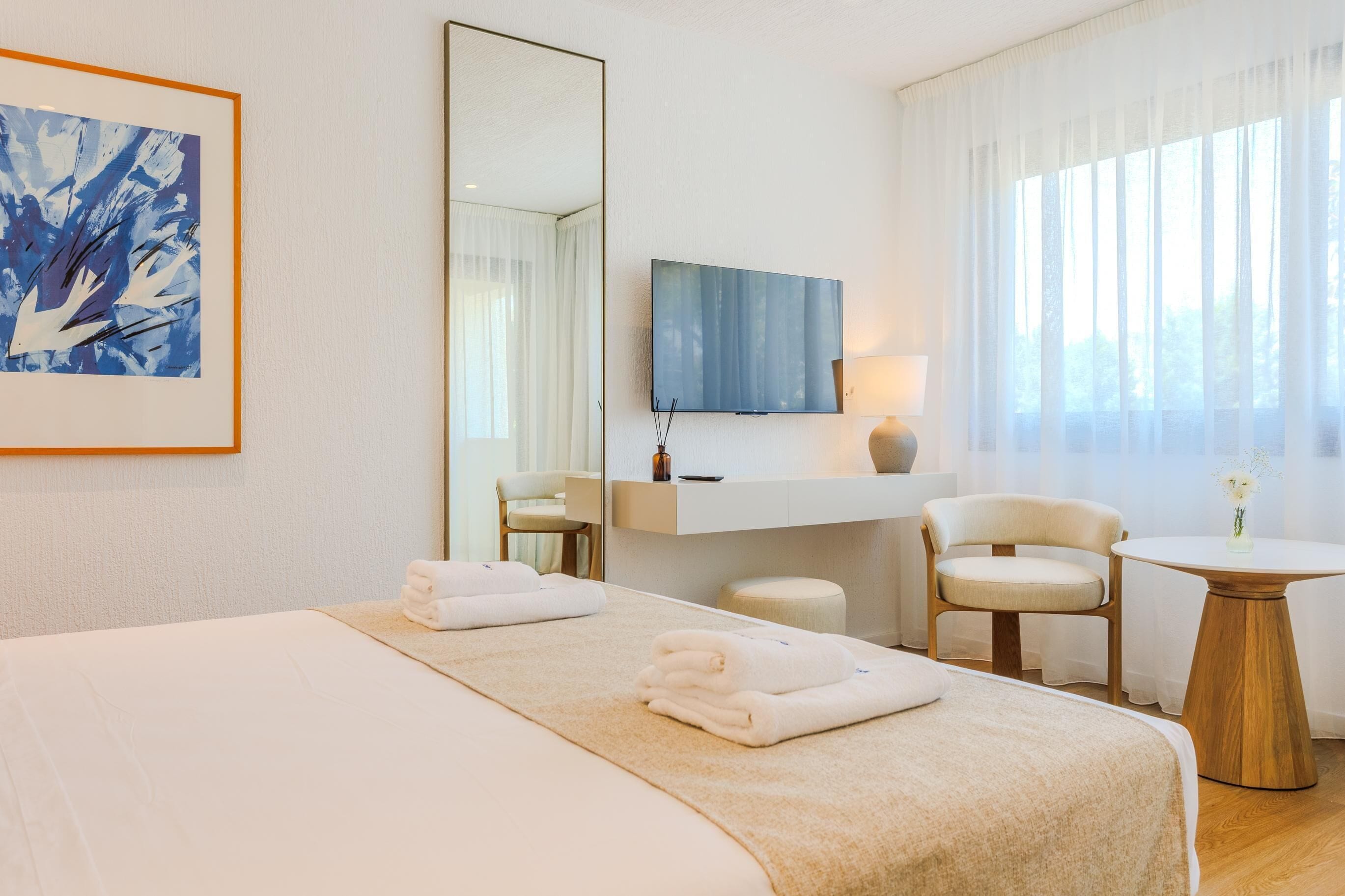 A hotel room with a bed, a desk, a chair, a TV, and a painting on the wall.