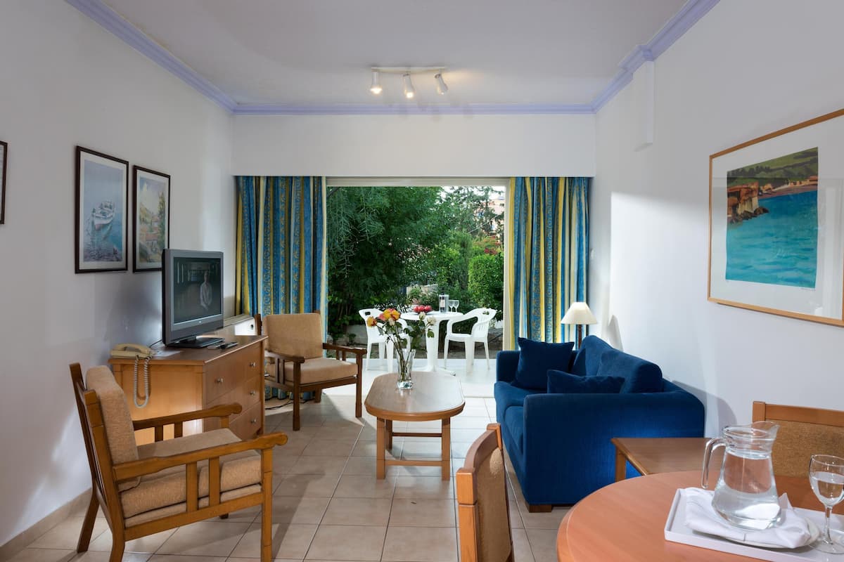 A hotel room with a blue sofa, wooden chairs, a small table with a pitcher and glass, a TV, and a balcony with a table and chairs.