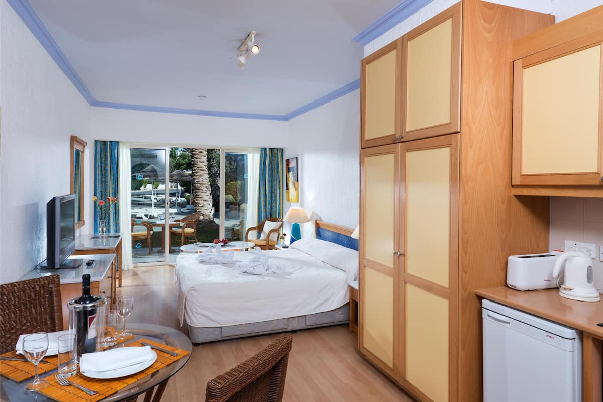 A hotel room with a bed, a dining table with chairs, a TV, a minibar, and a view of the outside.