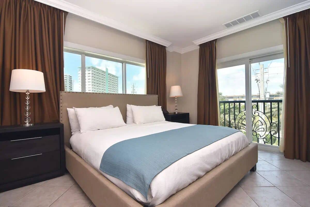 Ein Hotelzimmer mit einem großen Bett, zwei Nachttischen, einem Beistelltisch und einem Balkon mit Blick auf Gebäude.
