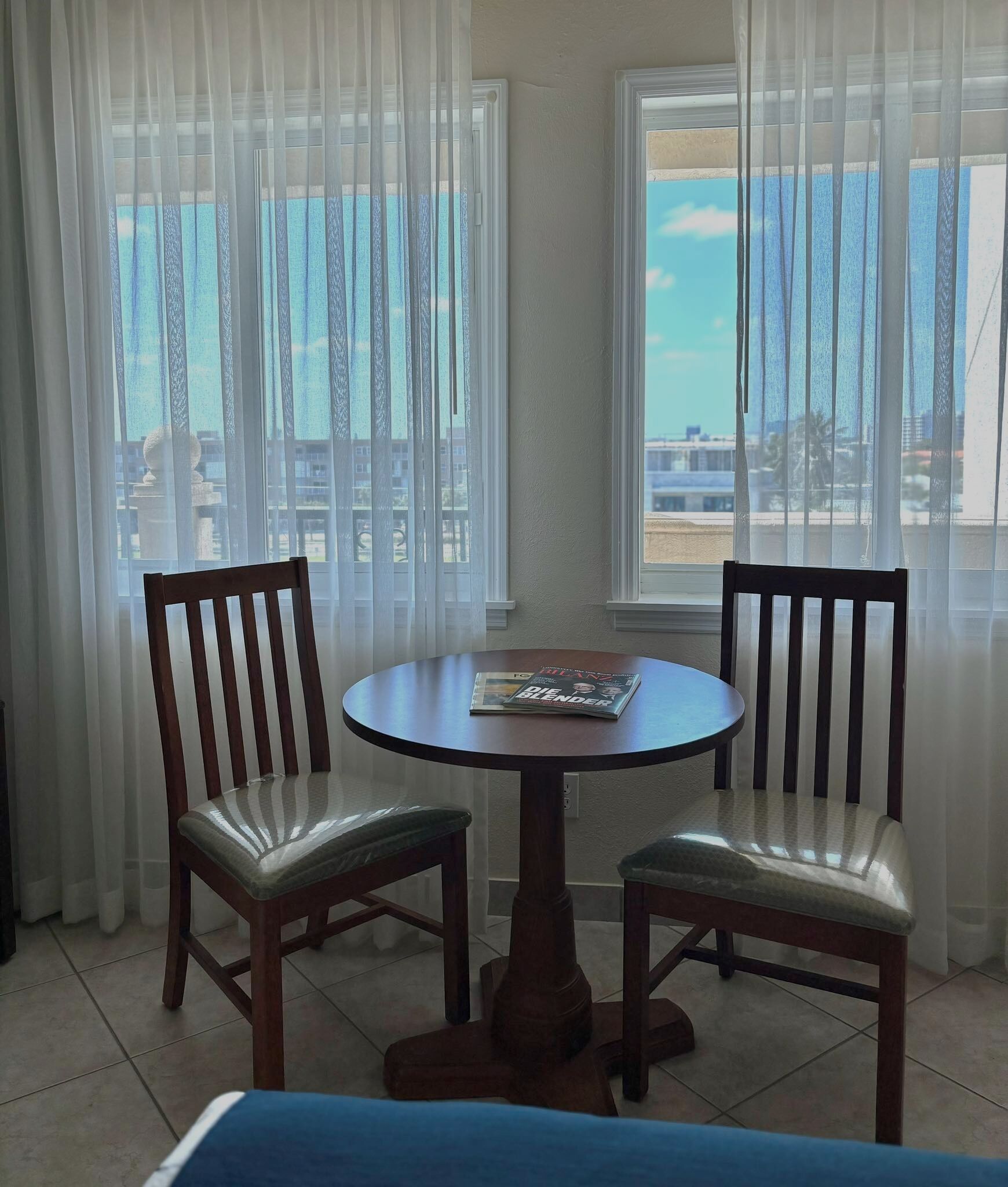 Un comedor con una mesa redonda y dos sillas de madera, una revista sobre la mesa, y vista al mar a través de cortinas translúcidas.
