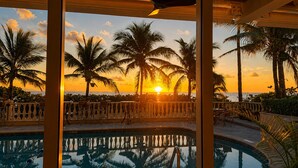 Piscina com vista para o pôr do sol, palmeiras e uma varanda com cadeiras.