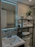 Une salle de bain moderne avec un grand miroir, un lavabo blanc, des toilettes et une étagère où sont rangées des serviettes et des articles de toilette.