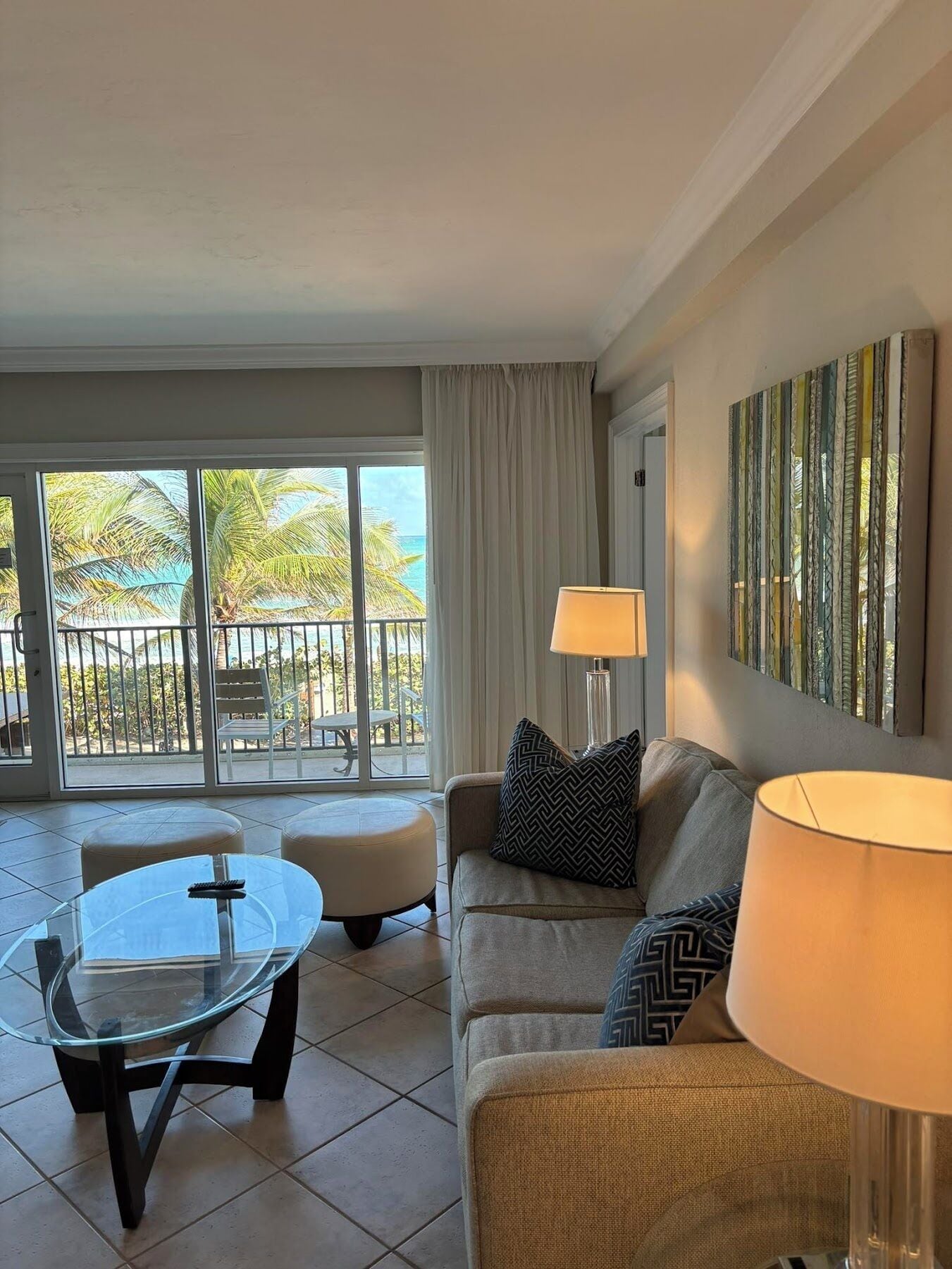 A living room with a glass table, a sofa, two lamps, and a view of the ocean through large windows.
