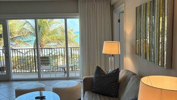 A living room with a glass table, a sofa, two lamps, and a view of the ocean through large windows.