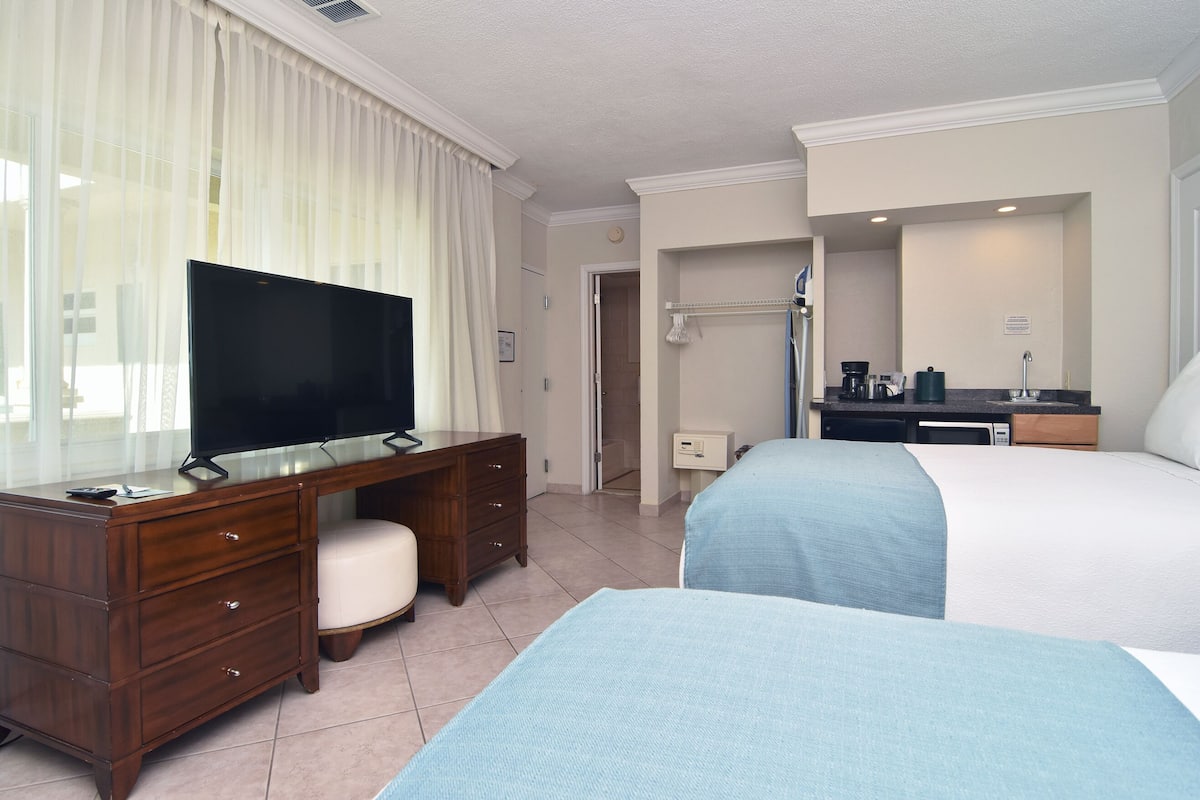 Ein Hotelzimmer mit Fernseher auf einem Holztisch, zwei Betten mit blauen Laken, einem Badezimmer mit Waschbecken und Spiegel sowie einer Duschkabine.
