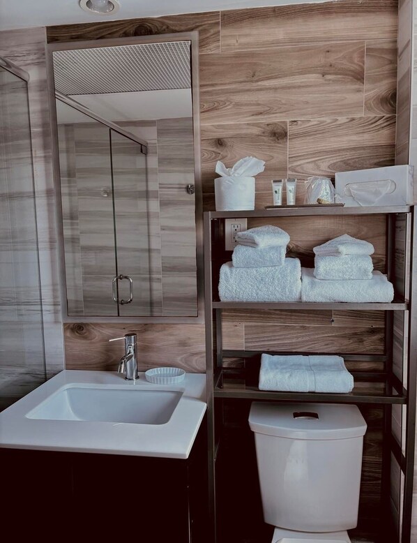 A modern bathroom with a wooden wall, a large mirror, a white sink, and neatly folded towels on shelves. - The Sea Lord (Lauderdale-by-the-Sea)