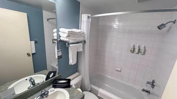 Un baño con lavamanos, espejo, inodoro y bañera. Hay toallas apiladas en un perchero y artículos de tocador en la pared.