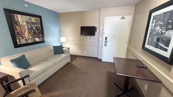 Quarto de hotel com sofá branco, uma pequena mesa, uma cadeira, televisão de tela plana na parede e porta com fechadura.
