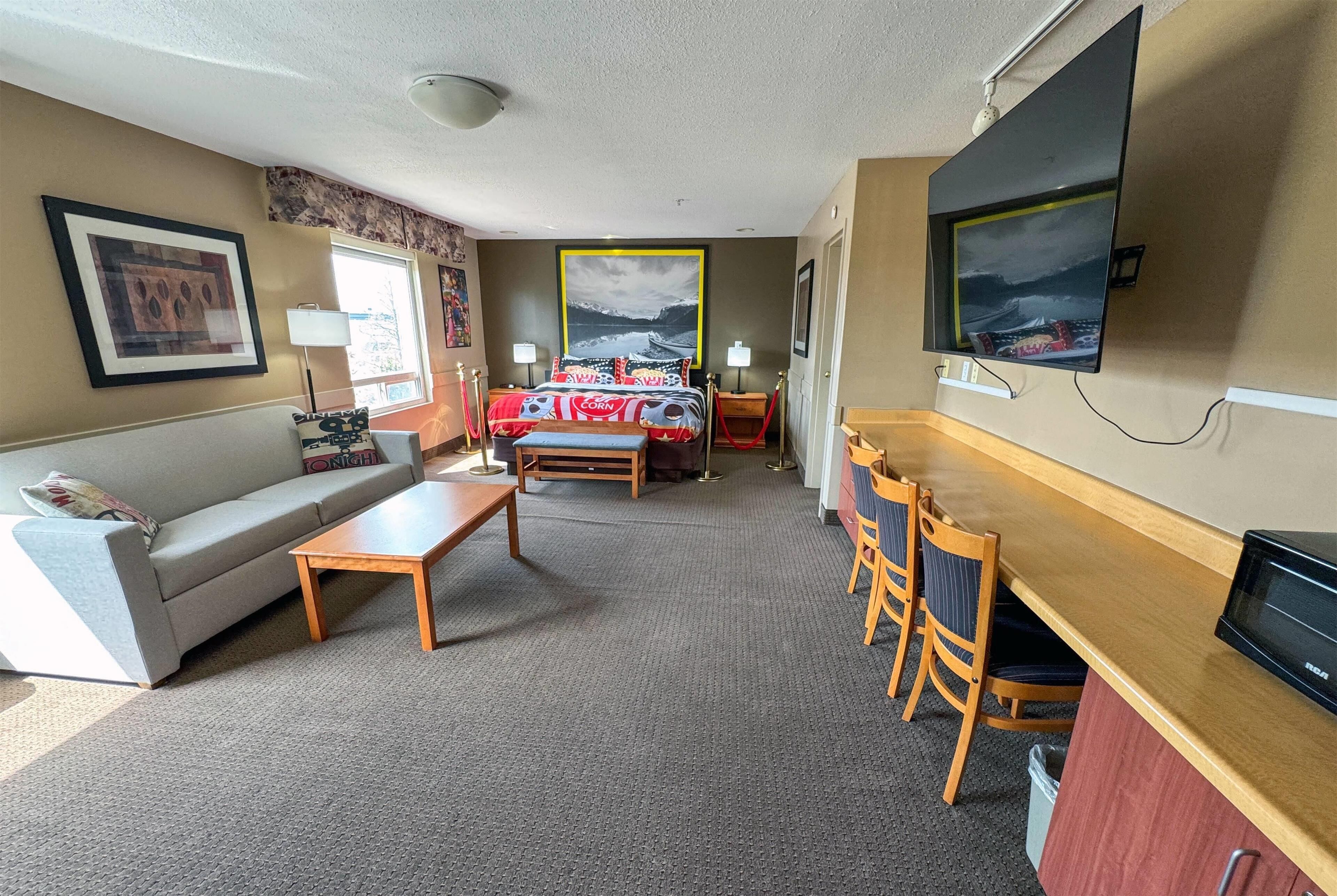 A hotel room with a sofa, a small table, a desk with chairs, a bed with a red and white bedspread, a TV mounted on the wall, and a framed picture above the bed.