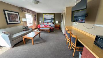 A hotel room with a sofa, a small table, a desk with chairs, a bed with a red and white bedspread, a TV mounted on the wall, and a framed picture above the bed.