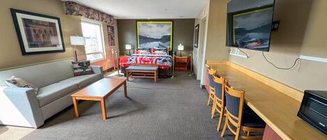 A hotel room with a sofa, a small table, a desk with chairs, a bed with a red and white bedspread, a TV mounted on the wall, and a framed picture above the bed.