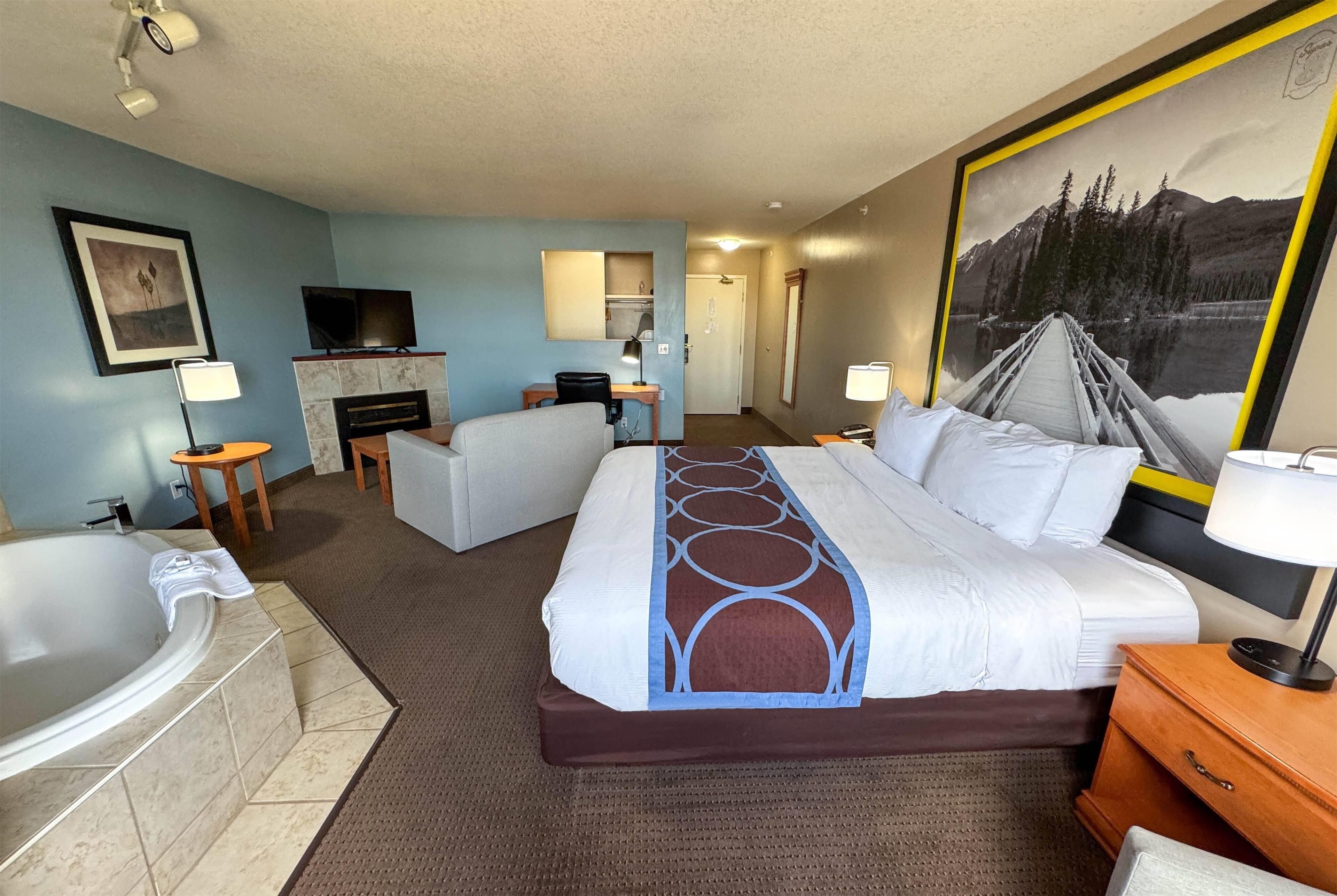 A hotel room with a large bed, a sitting area with a chair and a table, a TV, a fireplace, a bathroom with a bathtub, and a large framed picture on the wall.