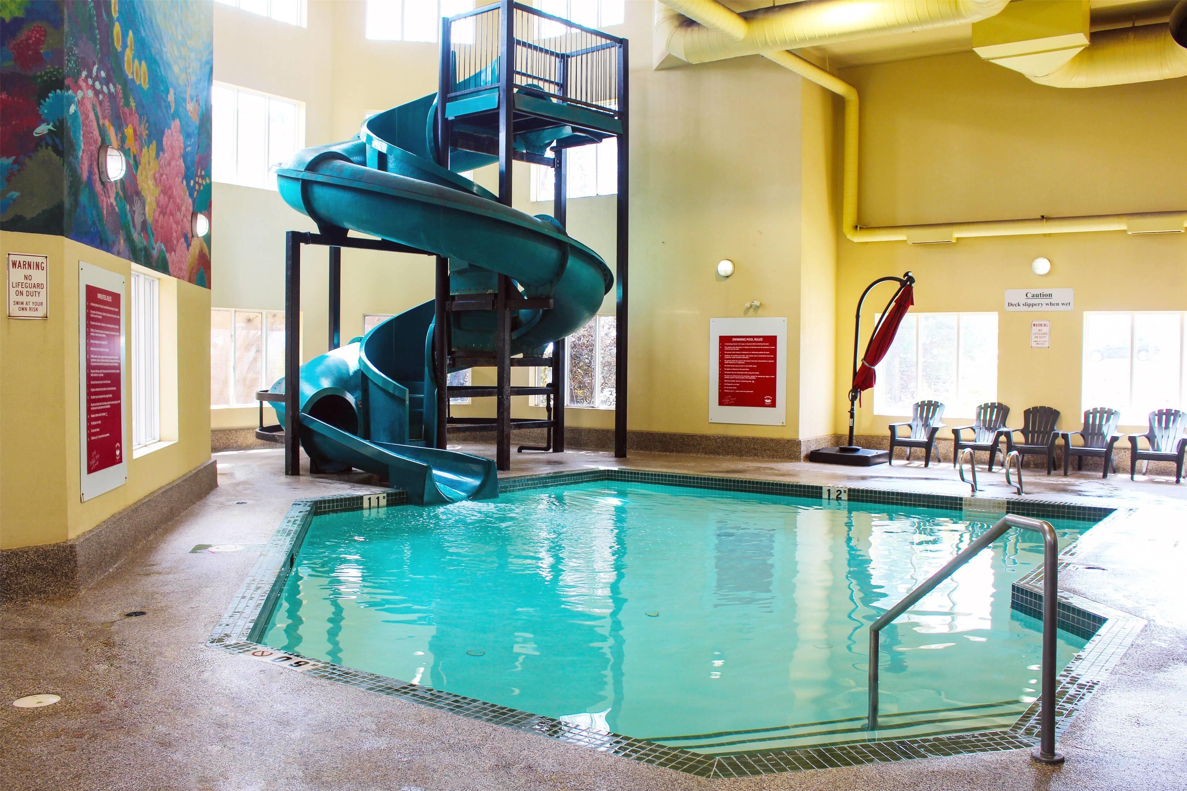 An indoor swimming pool with a blue spiral slide, safety signs, and a mural on the wall.