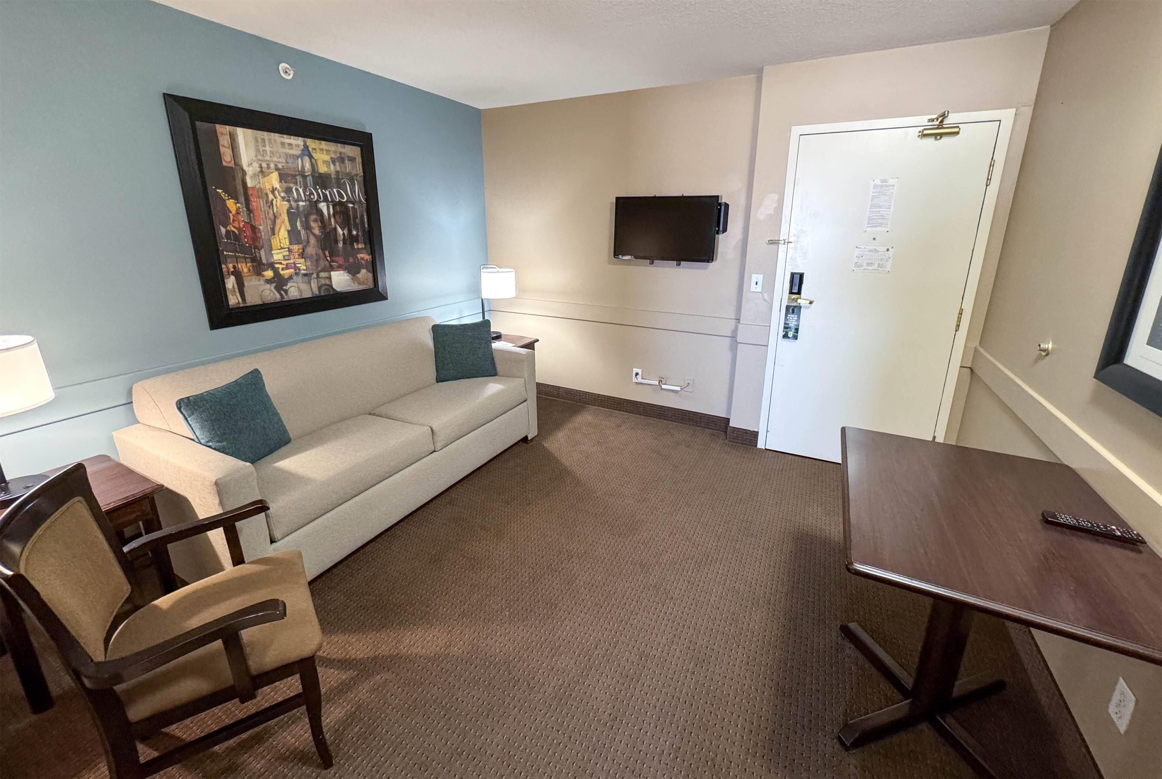 A hotel room with a sofa, a chair, a small table, a TV mounted on the wall, and a door with a lock.
