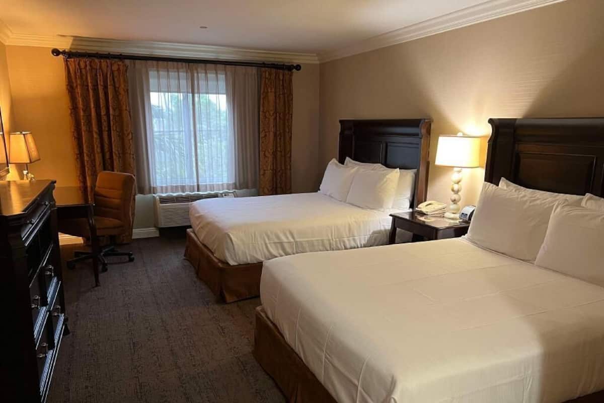 Hotellihuone, jossa on kaksi sänkyä, työpöytä, tuoli, lamppu ja ikkuna, jossa on verhot.