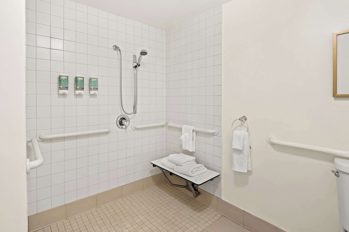 A bathroom with white tiled walls, a shower with a handheld showerhead, a grab bar, a bench with towels, and a toilet with a grab bar.