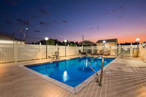 Una piscina con cerca blanca, sillas de descanso y un atardecer al fondo.