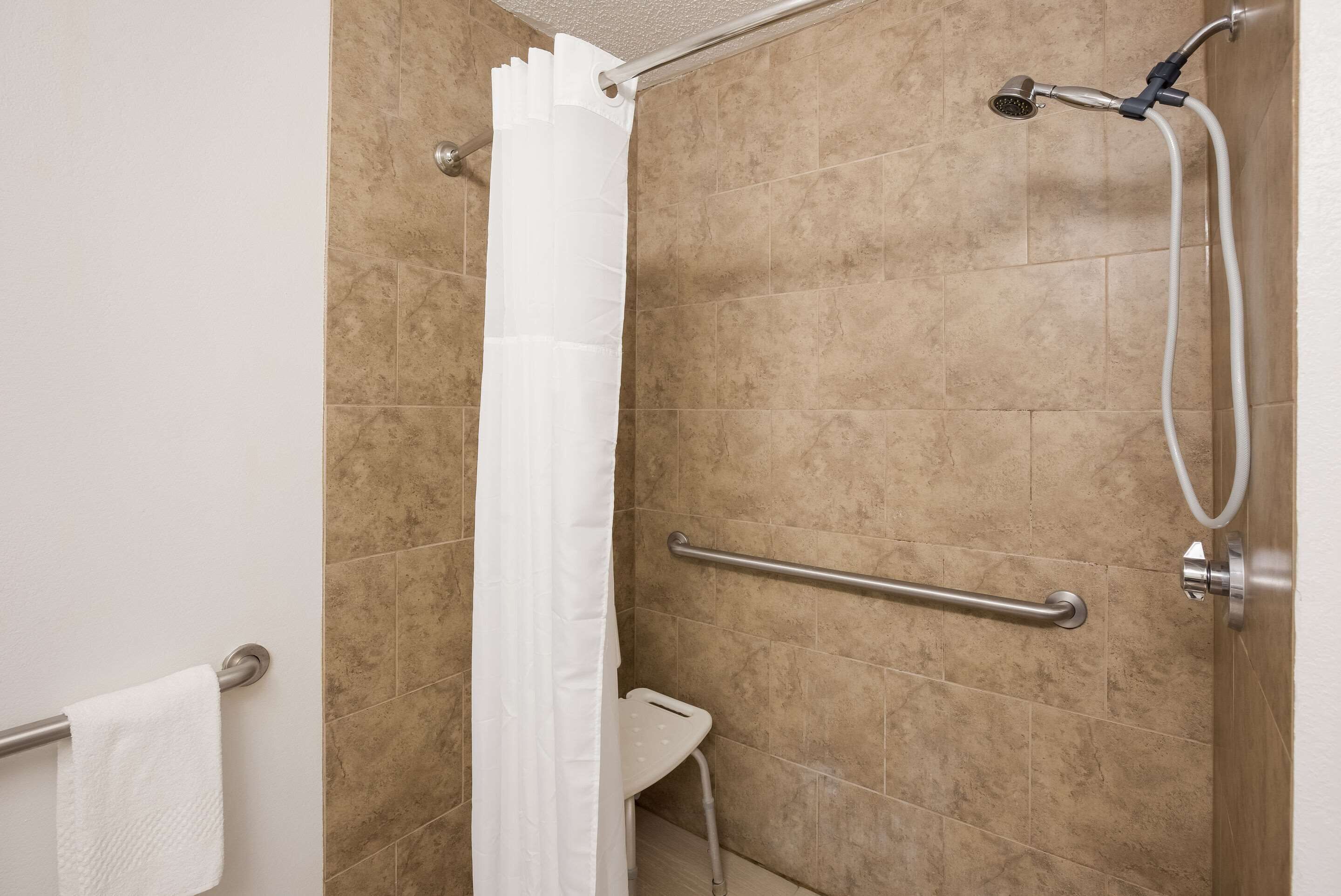 A shower area with beige tiles, a white shower curtain, a handheld showerhead, and a grab bar.