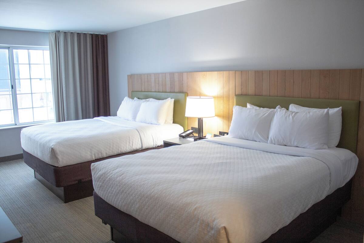 A hotel room with two beds, a wooden headboard, a bedside lamp, and a window with curtains.
