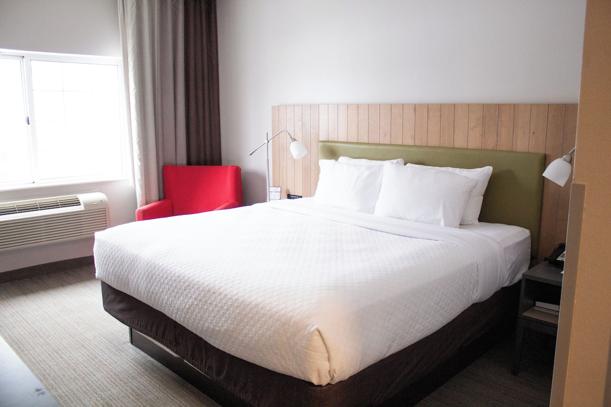 A hotel room with a large bed, a red chair, a bedside table with a lamp, and a window with curtains.