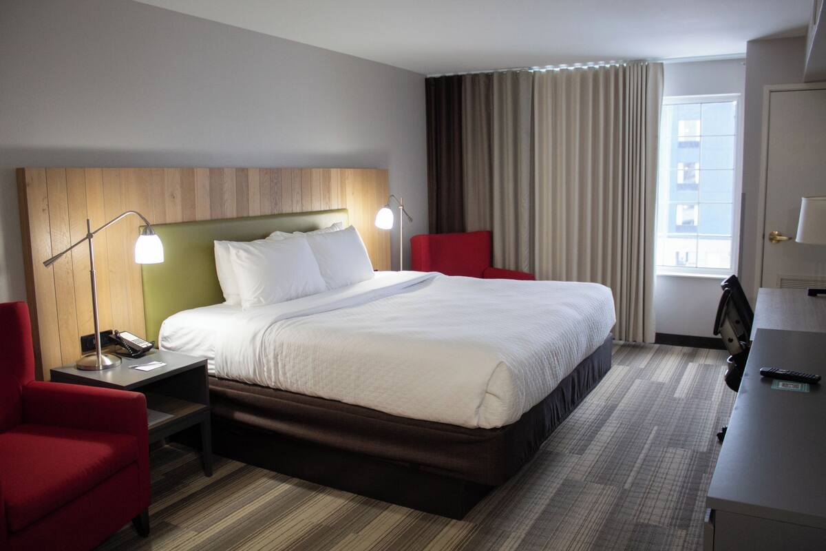 A hotel room with a large bed, a desk, a chair, and a nightstand with a lamp.