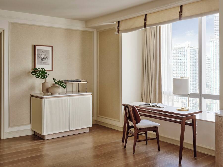 Habitación de hotel moderna con piso de madera, una mesa de comedor con una silla, un radiador, una planta y una ventana con cortinas.