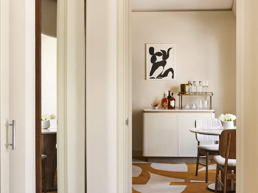 A hallway leading to a dining area with a white cabinet, a table with chairs, and a large mirror.