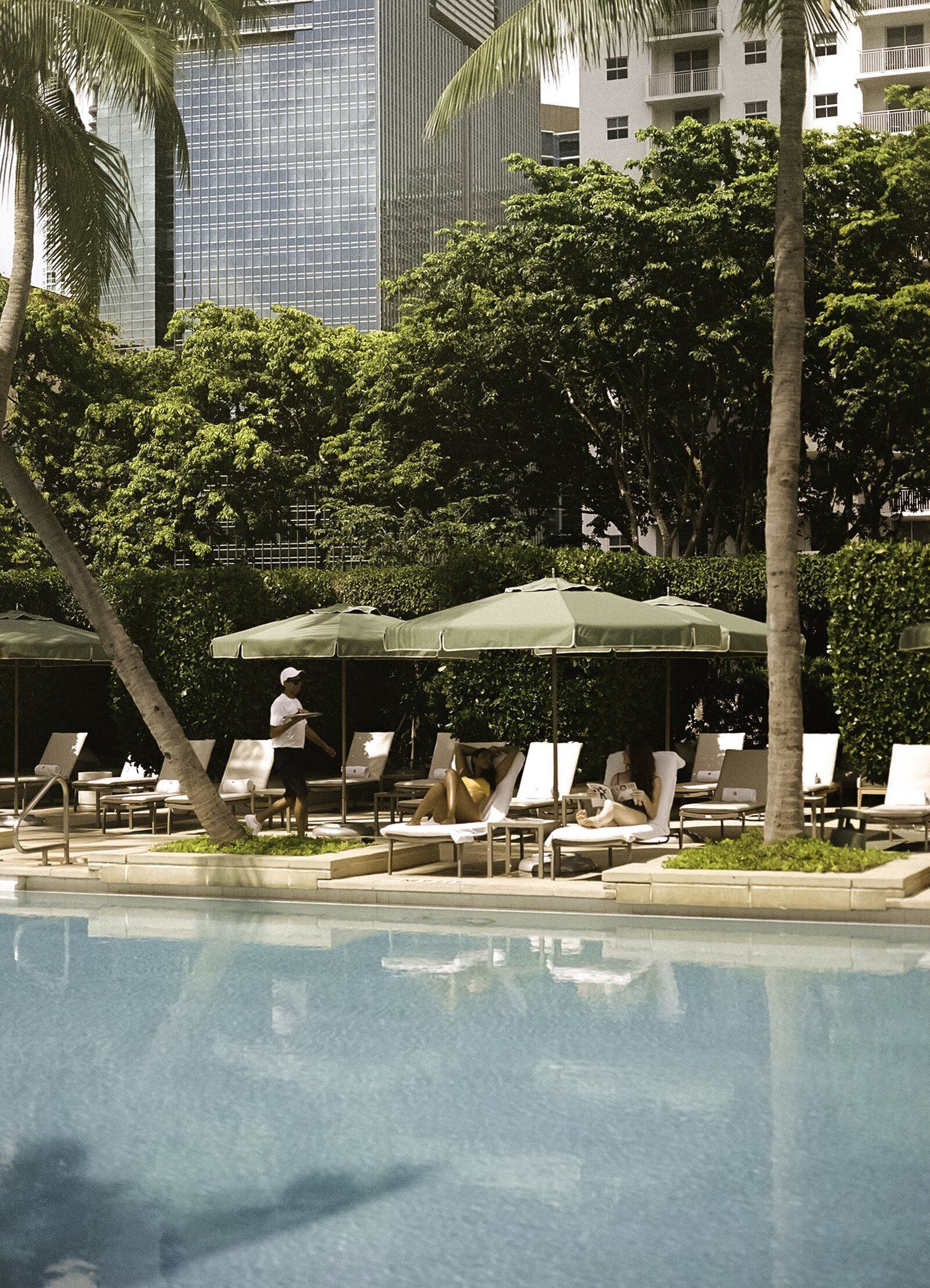 A poolside area with lounge chairs and umbrellas, surrounded by trees and a cityscape in the background.